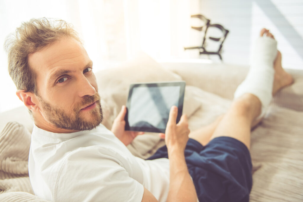 Mann mit Beinverband am Tablet zu Hause auf dem Sofa