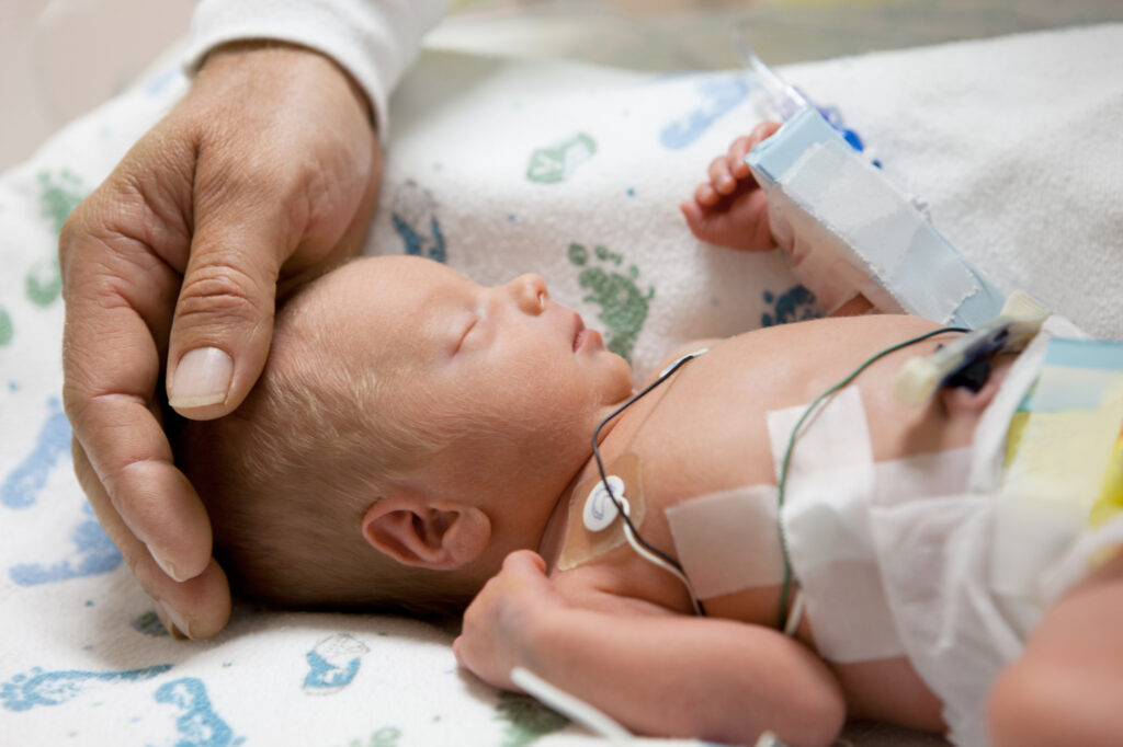 eine Hand streichelt den Kopf eines intensivmedizinisch versorgten Säuglings im Kinderbett im Krankenhaus
