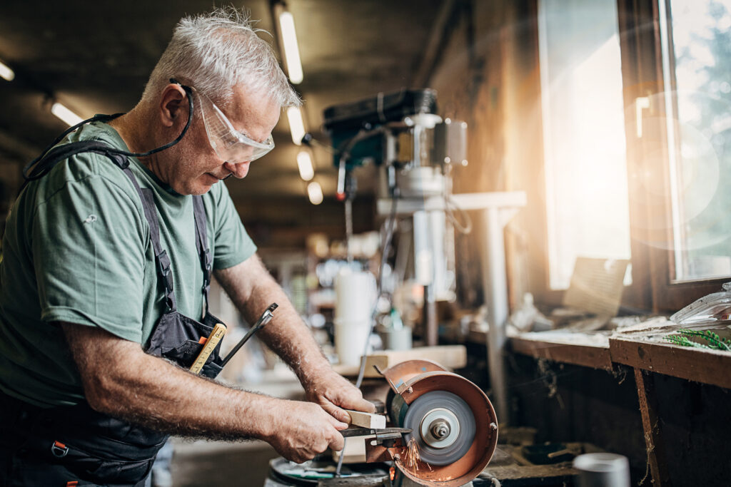 Älterer Mann steht in der Werkstatt beim Schleifen von Metall