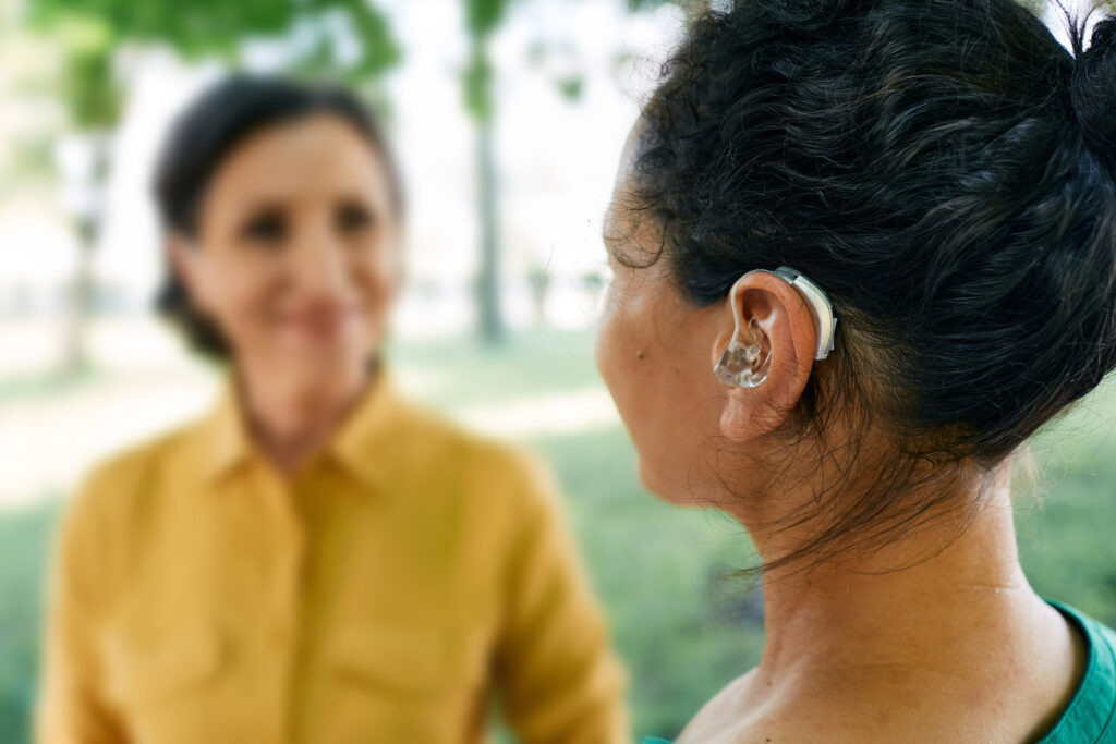 Erwachsene Frau mit Hörproblemen benutzt ein Hörgerät um sich mit einer Freundin im Park zu unterhalten.