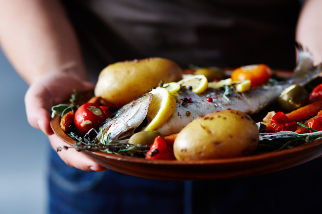 Nahaufnahme eines nicht erkennbaren Mannes, der einen Teller mit köstlichem Fisch hält, der mit Kartoffeln, Gemüse, Gewürzen und Zitrone gebacken wird