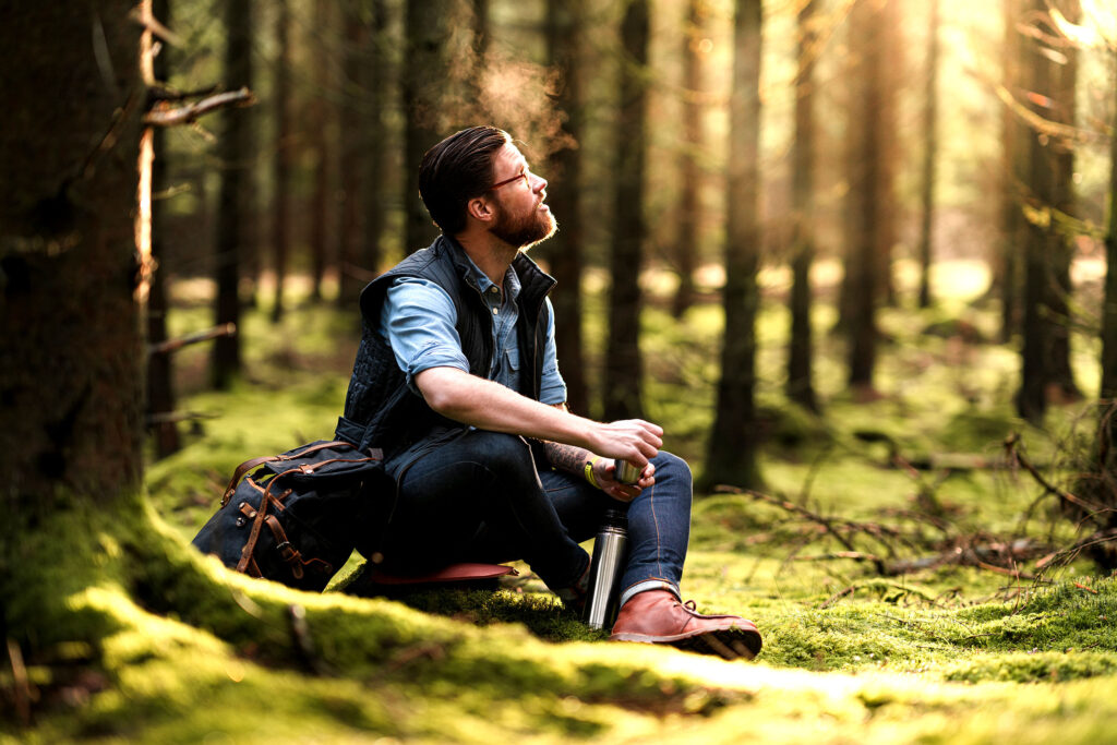 Mittelalter Mann sitzt nach einer Wanderung auf einer Lichtung im Wald und genießt sichtlich die Abgeschiedenheit und Ruhe bei einem Kaffe aus der Thermoskanne im Grünen