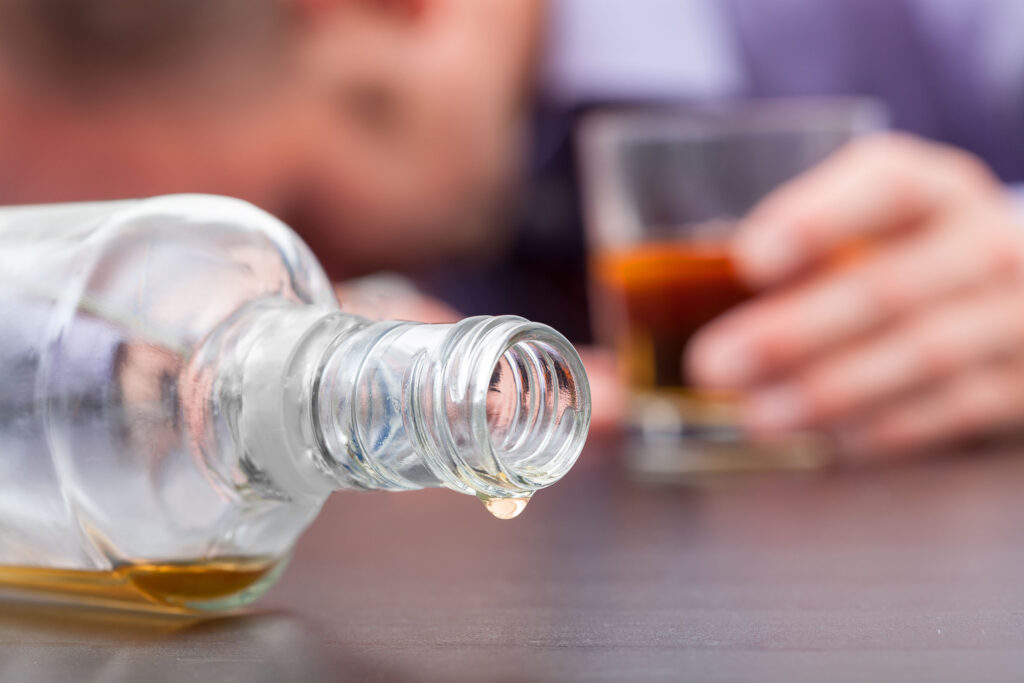 Symbolbild Alkoholmissbrauch: Bild zeit im Vordergrund eine leere liegende Schnapsflasche und unscharrf im Hintergrund einen offensischtlich alkoholisierten Mann, der ein übervolles Whiskeyglas in der Hand hält.
