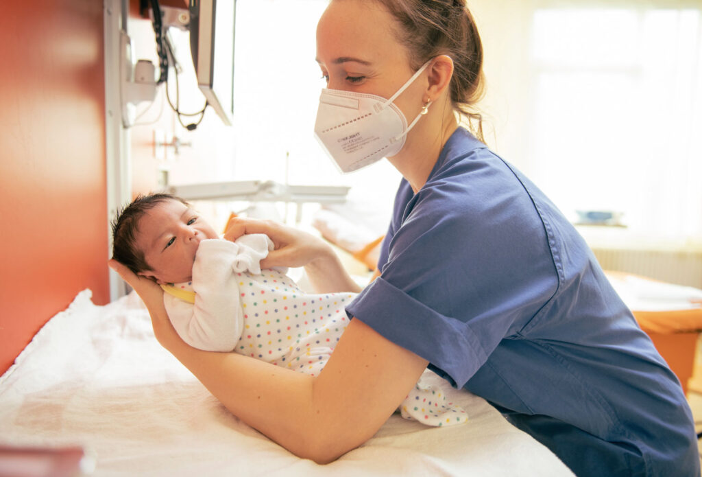 Kinderkrankenschwester hält behutsam ein Neugeborenes auf dem Arm und lächelt.