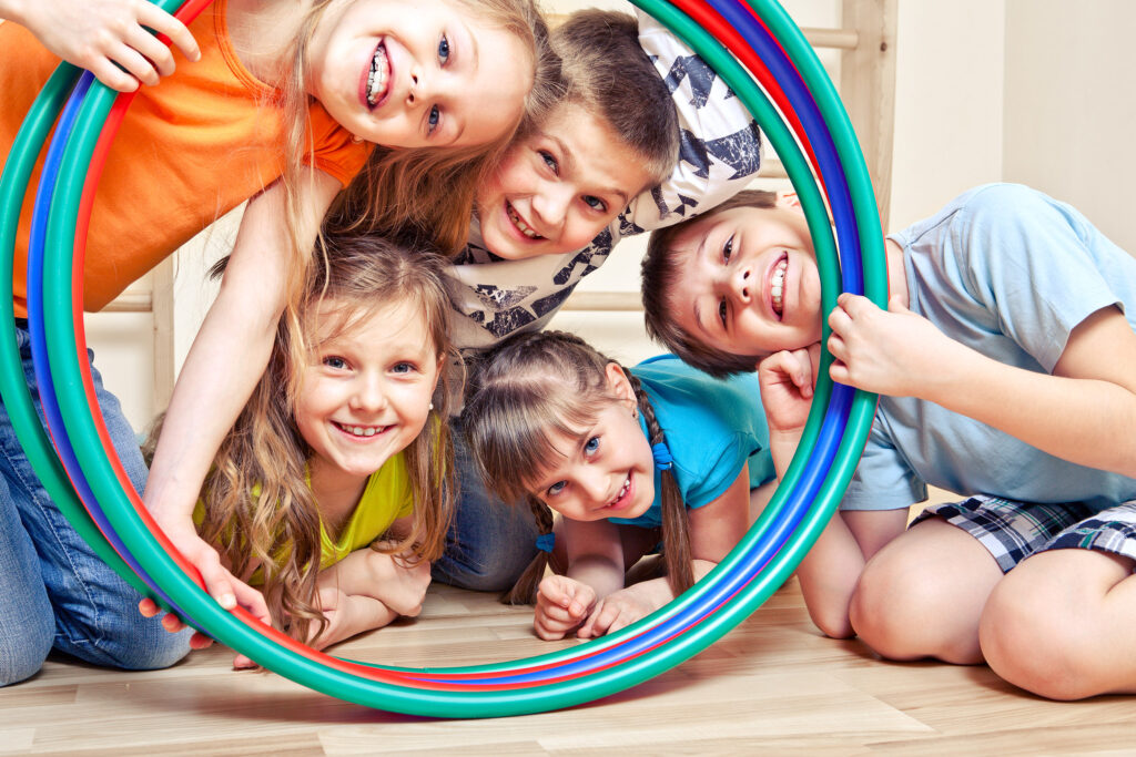 Rückengesundheit von Kindern: Fünf Kinder (männlich und weiblich) lächeln verscmitzt bzw. vergnügt durch mehrere Hulla Hoop Reifen