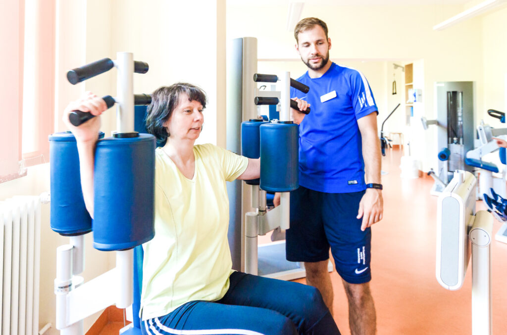 Eine Frau trainiert sitzend ihre Brustmuskularur am Trainingsgerät nach der Anweisung des Physiotherapeuten.