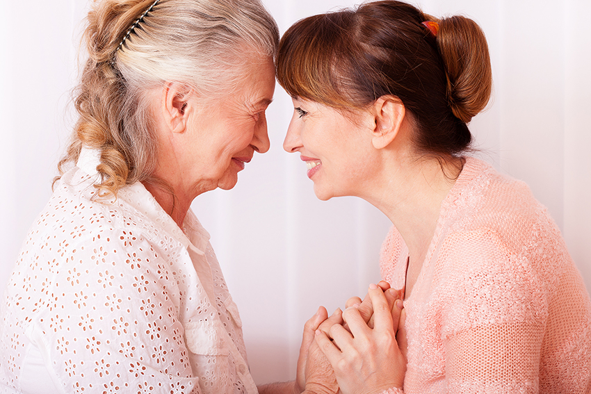 Alte Frau und Frau mittleren Alters stehen Stirn an Stirn, lächeln sich herzlich an. Gleichzeitig halten sich die beiden gegenseitig an den Händen.