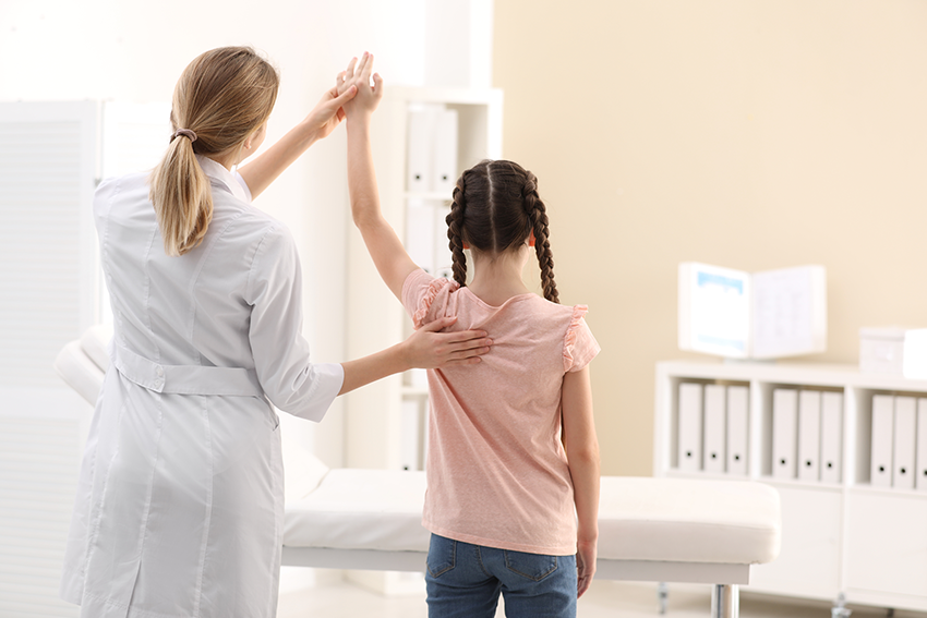 Eine Orthopädin steht neben einem Kind. Sie untersucht anhand einer Bewegung des Arms den Rücken des Kindes. Beide stehen mit dem Rücken zur Kamera in einem Untersuchungszimmer
