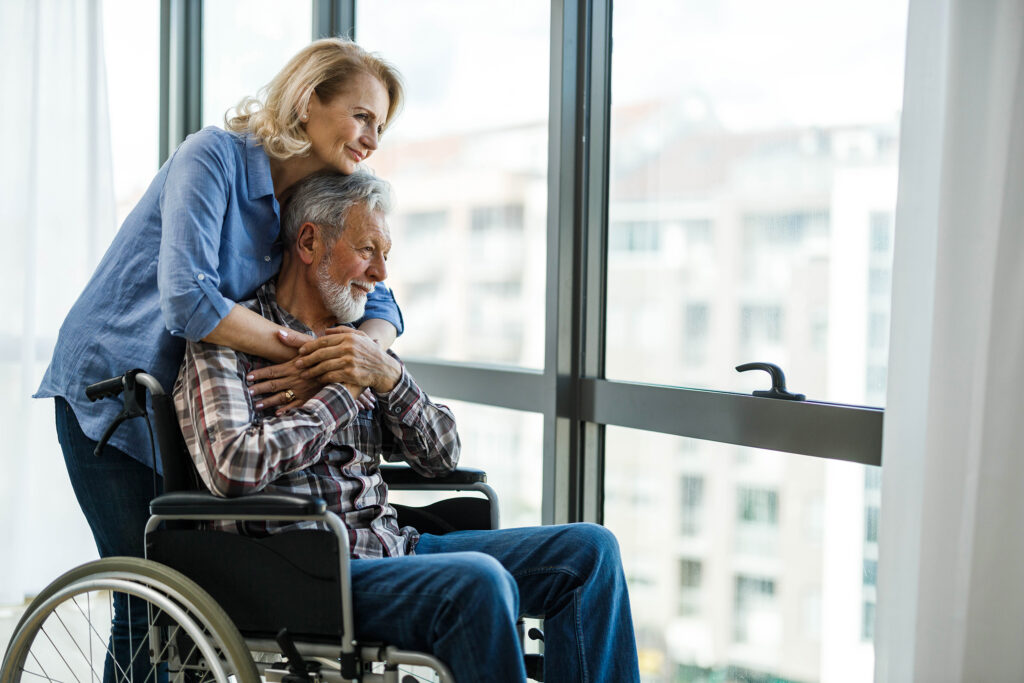 Eine ältere Frau und ein im Rollstuhl sitzender Mann blicken lächelnd aus dem Fenster ihrer Wohnung. Sie umarmt ihn von hinten, seine Hände ruhen auf ihren.